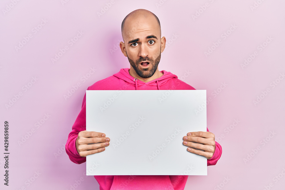 Young bald man holding blank empty banner in shock face, looking skeptical and sarcastic, surprised with open mouth Young bald man holding blank empty banner in shock face, looking skeptical and sarcastic, surprised with open mouth