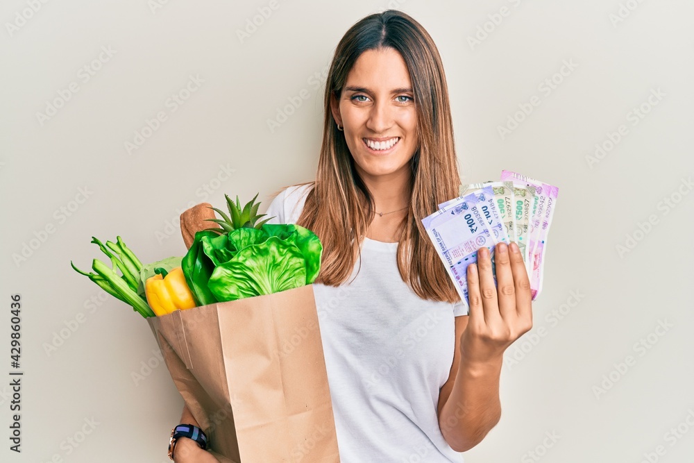 Brunette young woman holding groceries and indian rupee banknotes smiling with a happy and cool smile on face. showing teeth. Brunette young woman holding groceries and indian rupee banknotes smiling with a happy and cool smile on face. showing teeth.