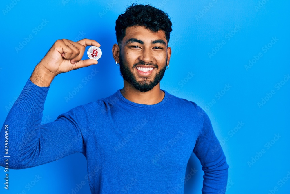 Arab man with beard holding virtual currency bitcoin looking positive and happy standing and smiling with a confident smile showing teeth Arab man with beard holding virtual currency bitcoin looking positive and happy standing and smiling with a confident smile showing teeth