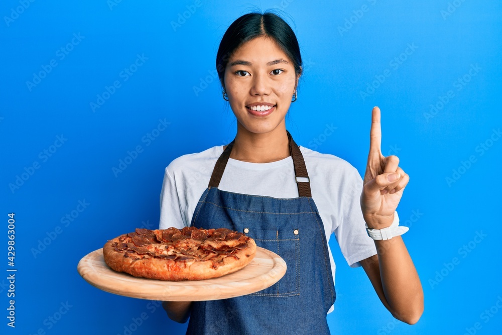 Young chinese woman wearing waiter apron holding italian pizza smiling with an idea or question pointing finger with happy face, number one Young chinese woman wearing waiter apron holding italian pizza smiling with an idea or question pointing finger with happy face, number one