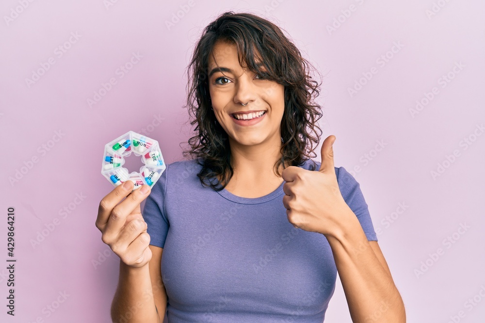 Young hispanic woman holding pill organizer smiling happy and positive, thumb up doing excellent and approval sign Young hispanic woman holding pill organizer smiling happy and positive, thumb up doing excellent and approval sign