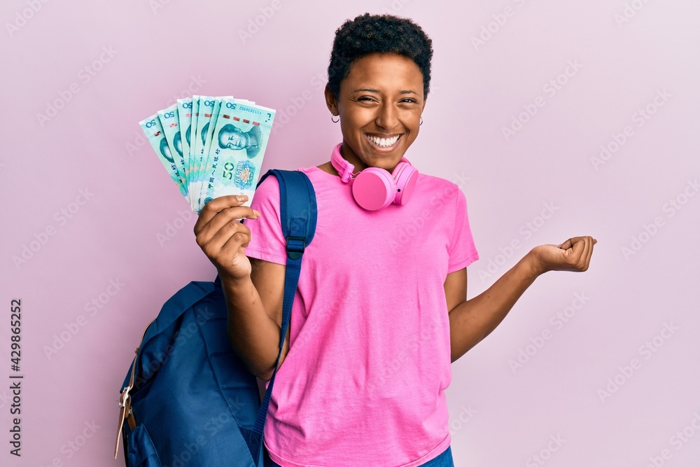 Young african american girl wearing student backpack holding chinese yuan banknotes screaming proud, celebrating victory and success very excited with raised arm Young african american girl wearing student backpack holding chinese yuan banknotes screaming proud, celebrating victory and success very excited with raised arm