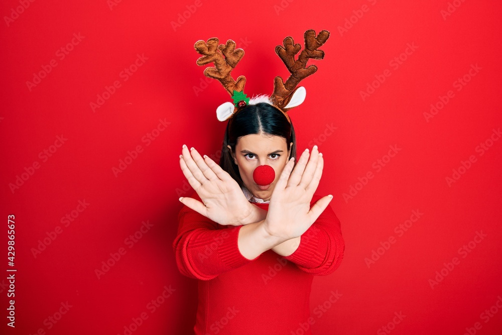 Young hispanic woman wearing deer christmas hat and red nose rejection expression crossing arms and palms doing negative sign, angry face