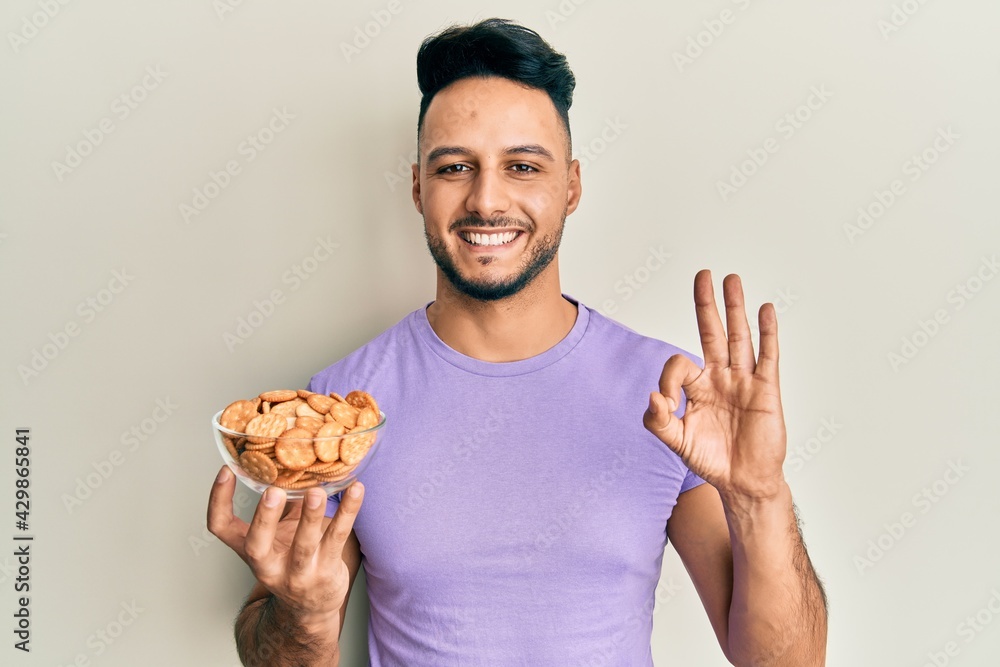 Young arab man holding bowl of salty crackers biscuits doing ok sign with fingers, smiling friendly gesturing excellent symbol Young arab man holding bowl of salty crackers biscuits doing ok sign with fingers, smiling friendly gesturing excellent symbol