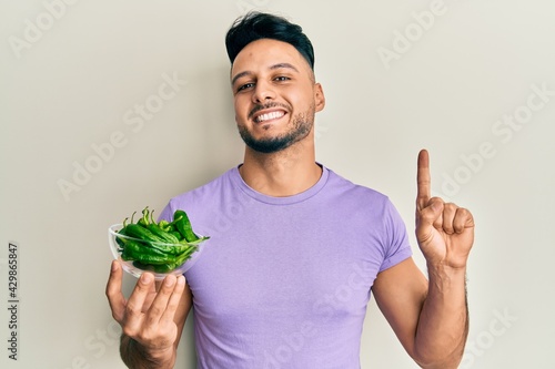 Young arab man holding bowl of green peppers smiling with an idea or question pointing finger with happy face, number one