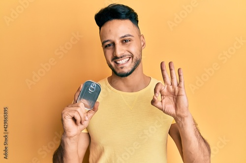 Young arab man holding canned food doing ok sign with fingers, smiling friendly gesturing excellent symbol