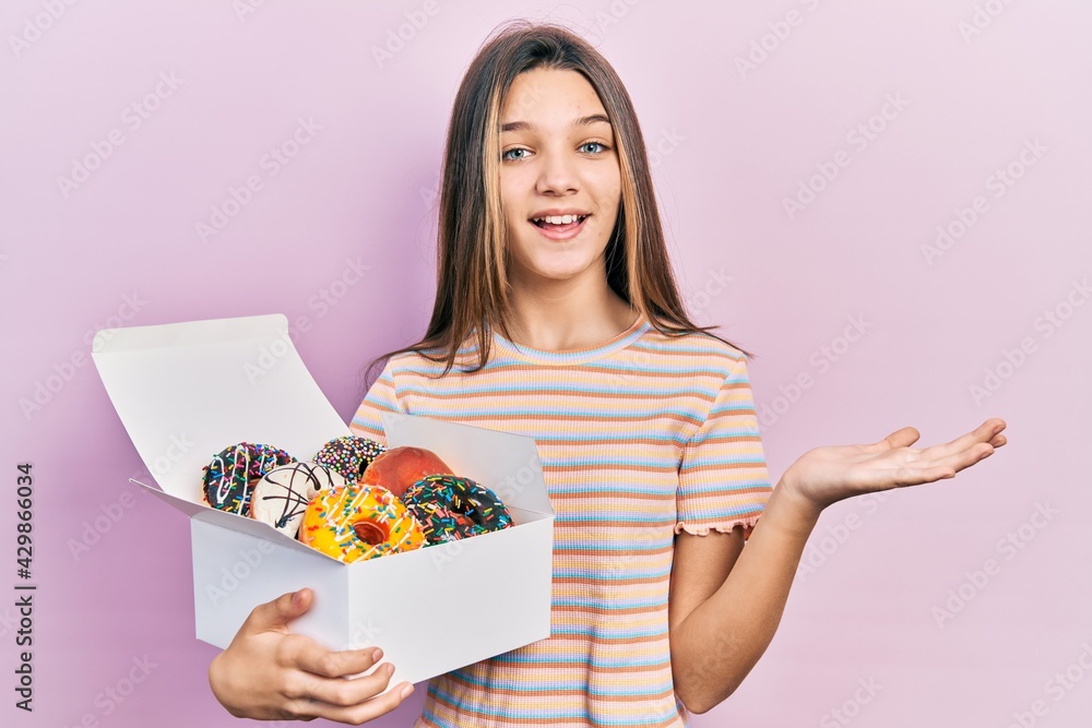 Young brunette girl holding tasty colorful doughnuts celebrating achievement with happy smile and winner expression with raised hand