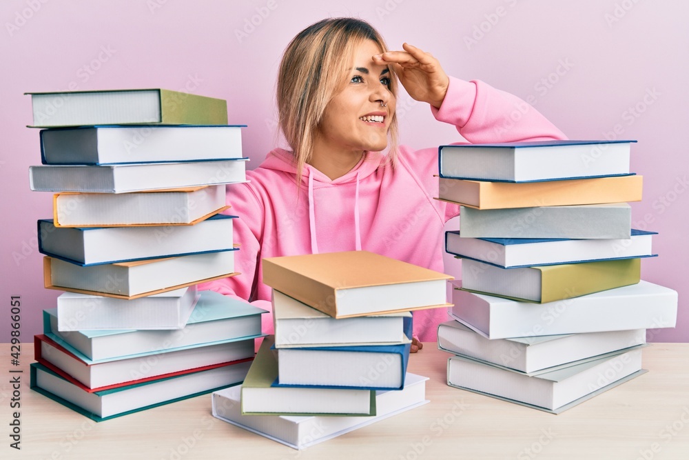 Young caucasian woman sitting on the table with books very happy and smiling looking far away with hand over head. searching concept. Young caucasian woman sitting on the table with books very happy and smiling looking far away with hand over head. searching concept.