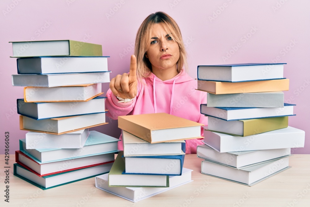 Young caucasian woman sitting on the table with books pointing with finger up and angry expression, showing no gesture Young caucasian woman sitting on the table with books pointing with finger up and angry expression, showing no gesture