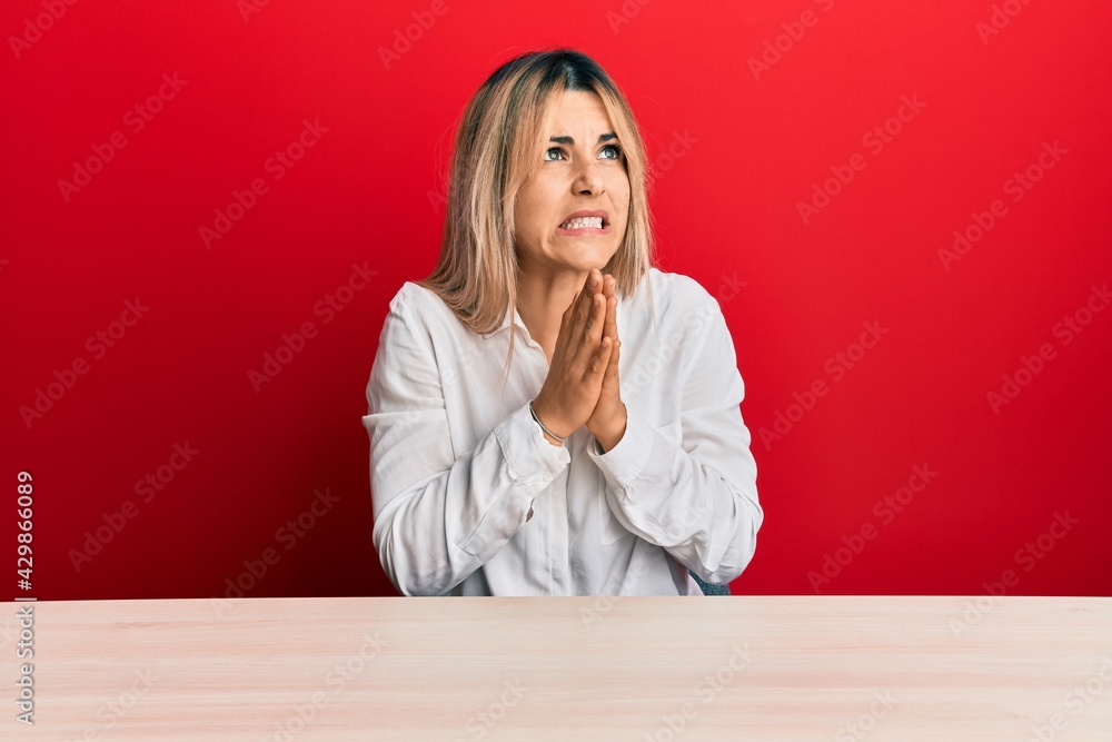 Young caucasian woman wearing casual clothes sitting on the table begging and praying with hands together with hope expression on face very emotional and worried. begging.