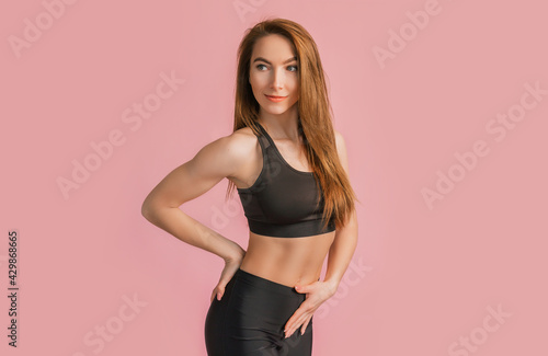 Fitness girl smiling in black sportswear on a pink background. Slim woman with a beautiful athletic body and tanned skin © Daria Lukoiko