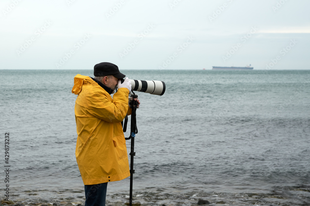 Landscape photography in bad weather. A man in a yellow raincoat takes photos from the sea coast with a telephoto lens supported by a monopod. Copy space.