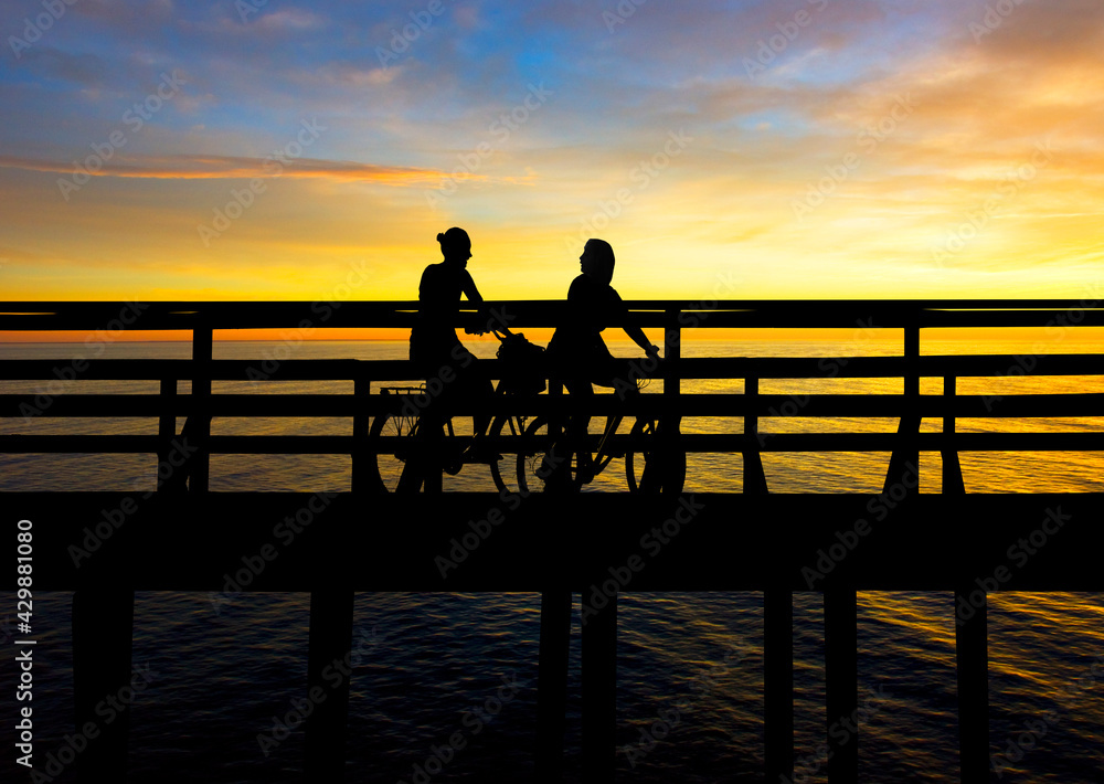 Two young women cycling on bridge at sunset
