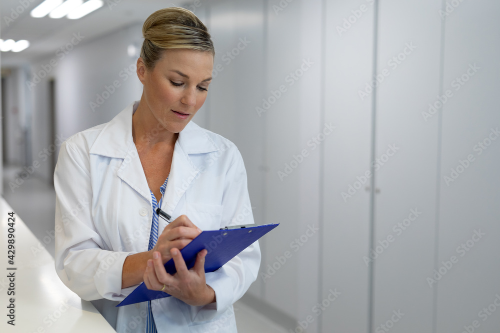 Caucasian female doctor standing in hospital corridor filling medical chart document Caucasian female doctor standing in hospital corridor filling medical chart document