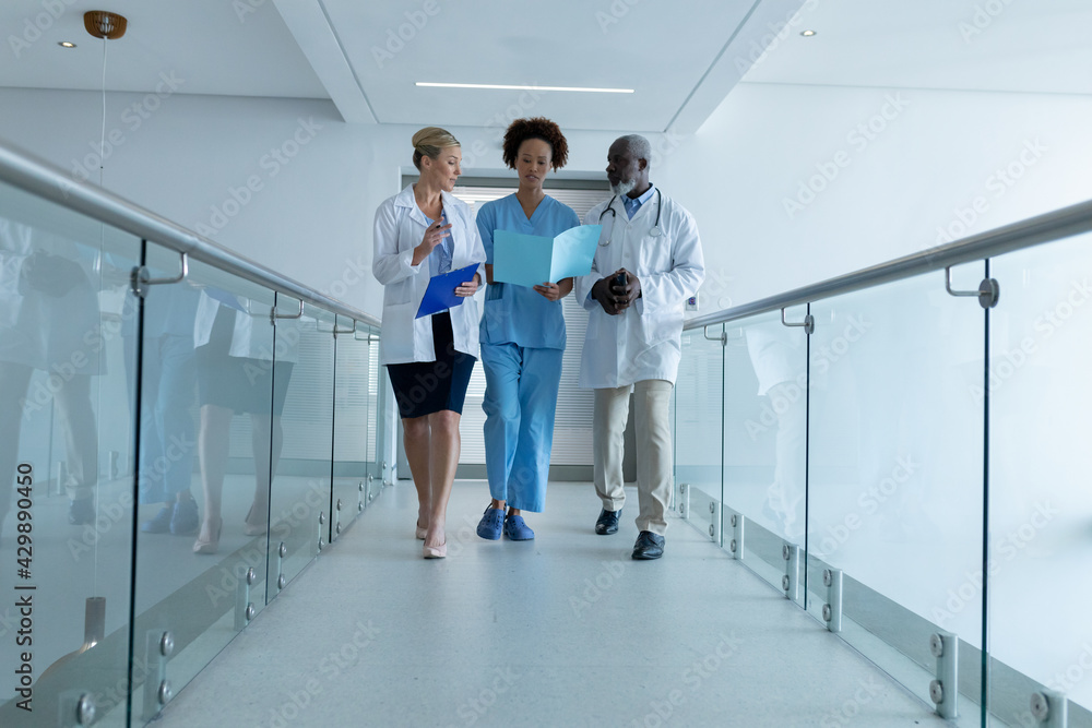 Three diverse male and female doctors walking through hospital corridor looking at document Three diverse male and female doctors walking through hospital corridor looking at document