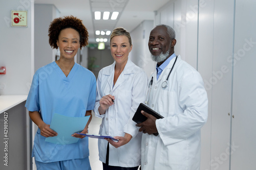 Portrait of three diverse male and female doctors standing in hospital corridor smiling to camera
