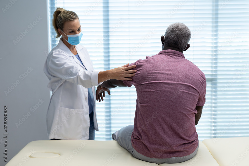 Obraz premium Caucasian female physiotherapist wearing mask stretching arm of african american male patient