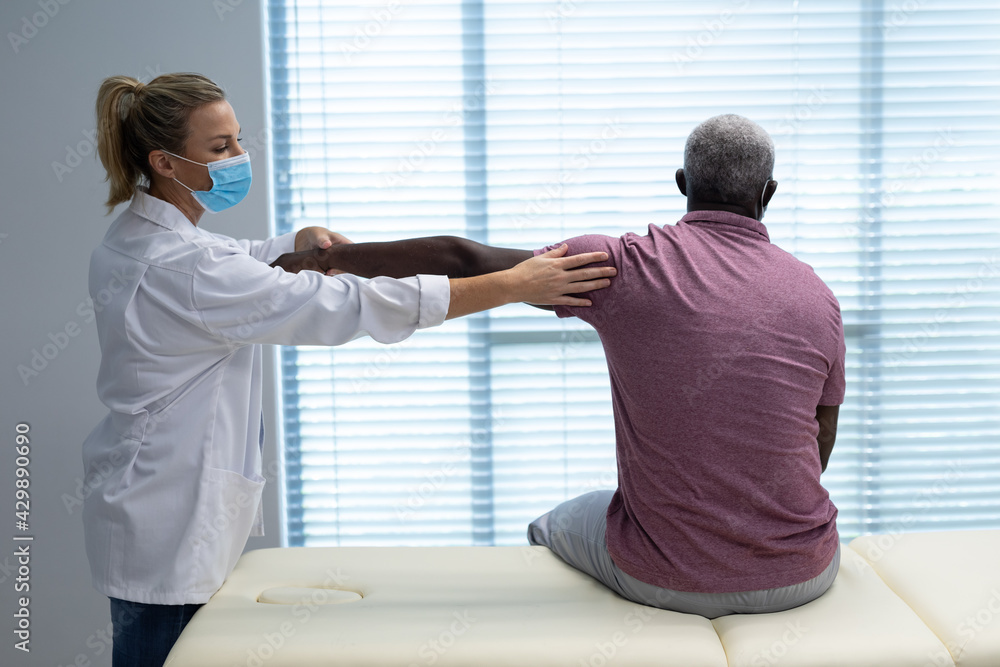 Caucasian female physiotherapist wearing mask stretching arm of african american male patient Caucasian female physiotherapist wearing mask stretching arm of african american male patient
