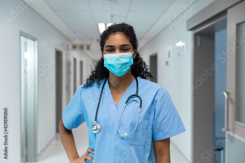 Portrait of mixed race female doctor wearing face mask standing in hospital corridor