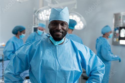 Smiling african american male surgeon with face mask and protective clothing in operating theatre