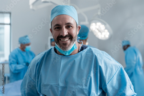 Smiling caucasian male surgeon with face mask and protective clothing in operating theatre