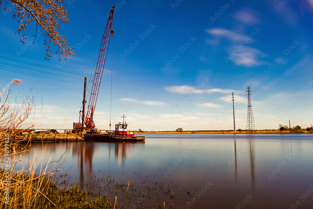 A fine art landscape image of a long exposure view of Nanticoke river on the coast of Vienna, Maryland near Chesapeake bay. A crane, a docked vintage cargo or tow boat and high voltage line tower seen