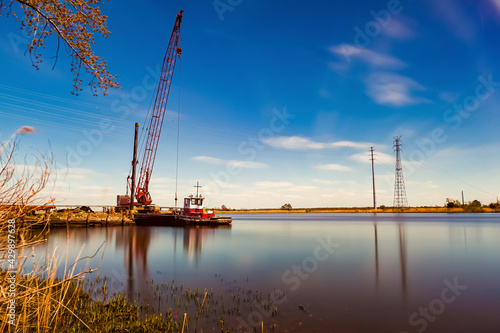 A fine art landscape image of a long exposure view of Nanticoke river on the coast of Vienna, Maryland near Chesapeake bay. A crane, a docked vintage cargo or tow boat and high voltage line tower seen