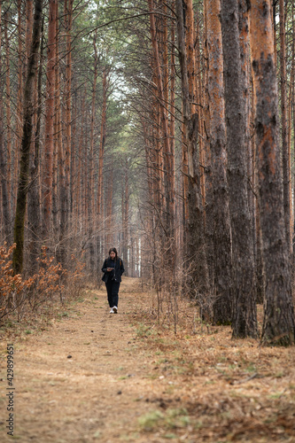  A girl with a phone in her hands and headphones in her ears is walking along a path in a pine forest.