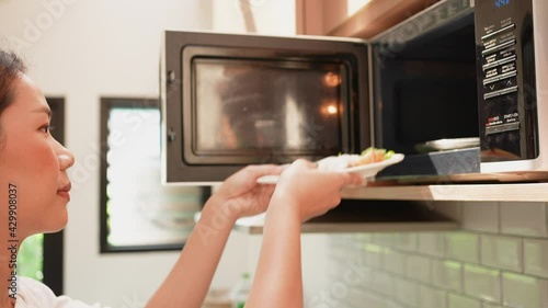 slow motion scene of asian young woman opening microwave oven to lay dish of food for defrost in the kitchen at home for comfortable lifestyle concept
