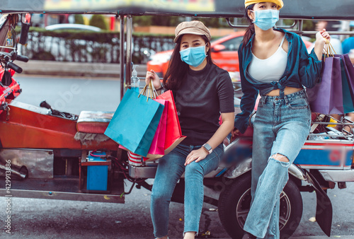 Asian woman wearing face mask. Happy woman with shopping bags.travel concept.group of happy friend are traveling.tourist holding shopping bag sitting in tuk tuk thai taxi with blurring town bangkok