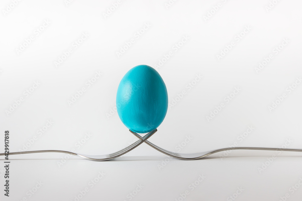 Easter composition with one blue colored egg on two crossed metallic forks on white background Easter composition with one blue colored egg on two crossed metallic forks on white background