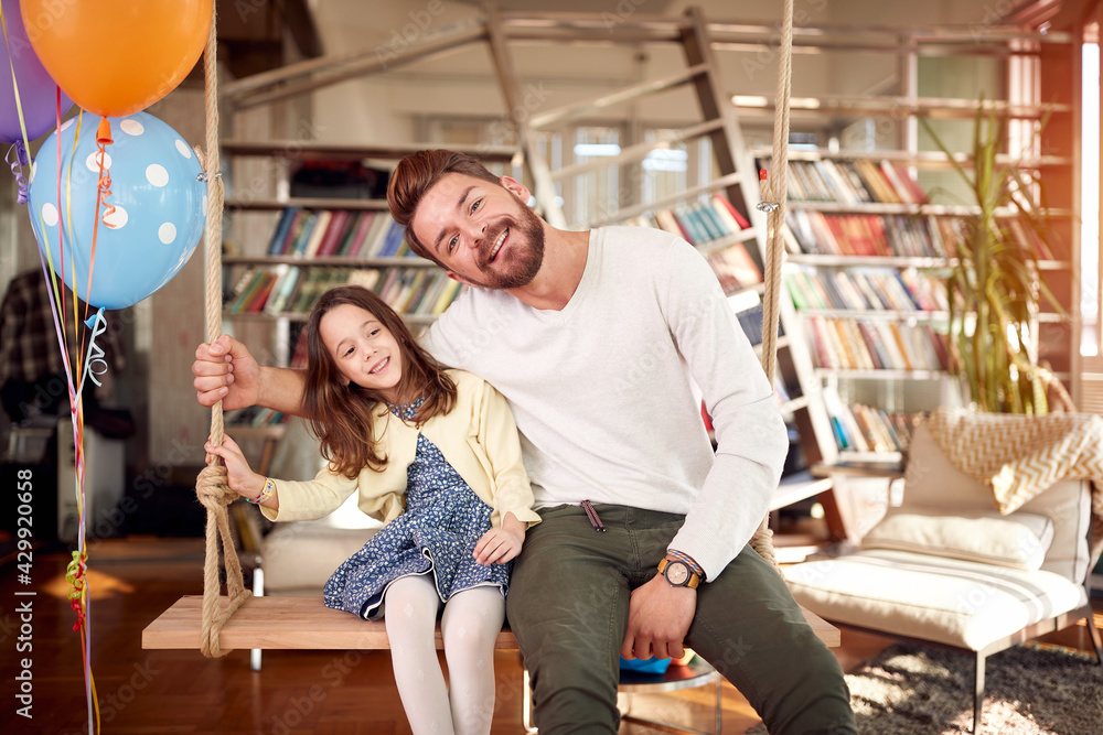 A happy Dad and his little daughter sitting on the swing at home and posing for a photo. Family, leisure, together