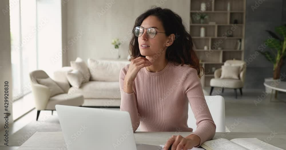 Thoughtful attractive woman working on laptop seated at desk in cozy living room. Businesswoman sit at workplace makes calculations in her head, text on computer having busy fruitful day at homeoffice