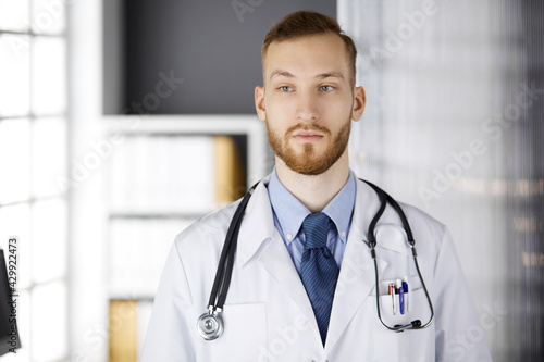 Red-bearded doctor standing straight in clinic near his working place. Portrait of physician. Medicine, healthcare