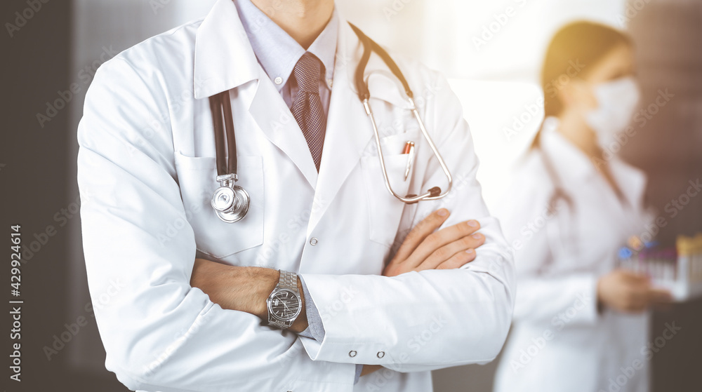 Unknown male doctor in protective mask standing with arms crossed in sunny clinic. Medicine concept during Coronavirus pandemic Unknown male doctor in protective mask standing with arms crossed in sunny clinic. Medicine concept during Coronavirus pandemic
