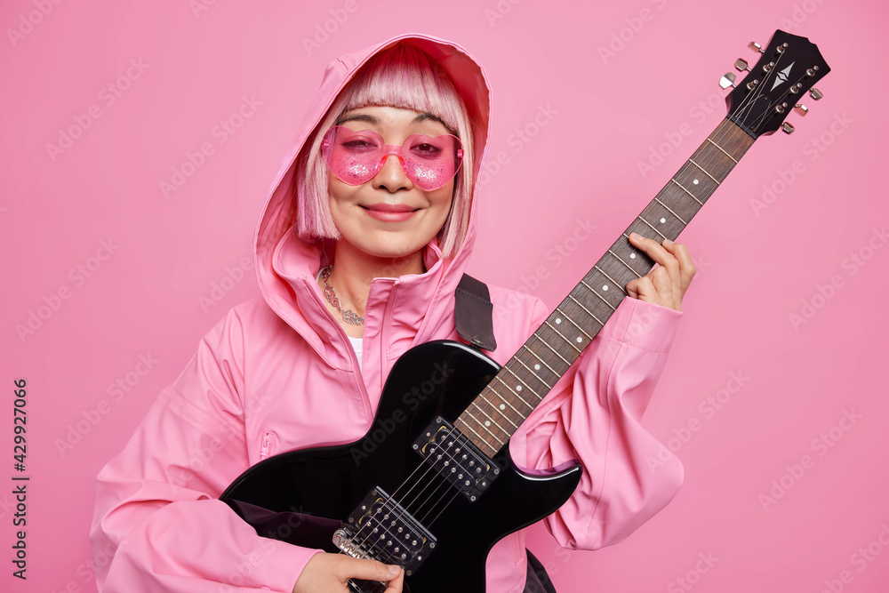 Fototapeta premium Photo of pleased fashionable female musician plays electric guitar smiles pleasantly wears heart shaped sunglasses anorak with hood poses against pink background. Monochrome shot. Music concept