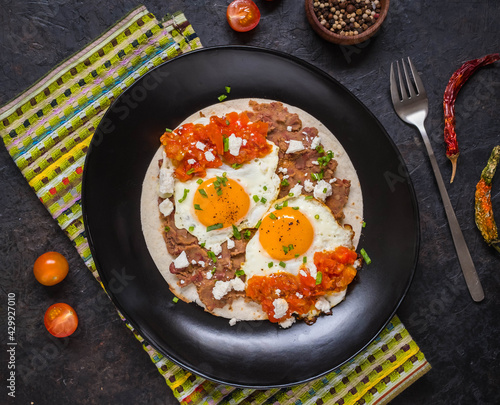 Bild auf Leinwand Huevos rancheros, Mexican fried egg on a wheat tortilla with tomato salsa, bean paste and feta cheese on a black plate on a light concrete background