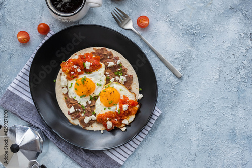 Huevos rancheros, Mexican fried egg on a wheat tortilla with tomato salsa, bean paste and feta cheese on a black plate on a light concrete background.