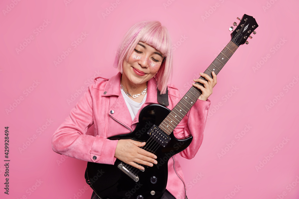 Pink haired pleased woman tilts head plays acoustic guitar enjoys favorite melody prepares for rock n roll concert wears fashionable clothes poses in studio against rosy background. Music concept
