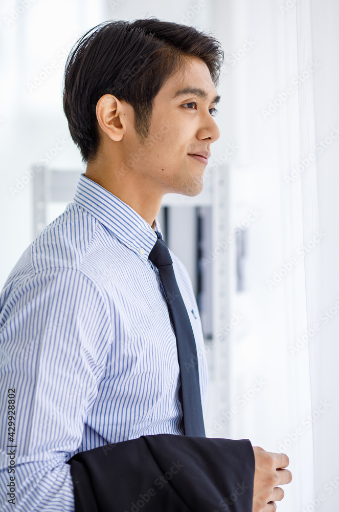 Portrait shot of Asian young happy successful businessman take black formal jacket uniform off hold on arms standing look outside thinking smiling in front white bookshelf background alone in library Portrait shot of Asian young happy successful businessman take black formal jacket uniform off hold on arms standing look outside thinking smiling in front white bookshelf background alone in library