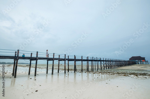 A woman dressed in light summer clothes walking on the Zanzibar island low tide sandy beach wooden pier. Careless vacation in the tropical countries concept image. Kiwengwa  Tanzania.