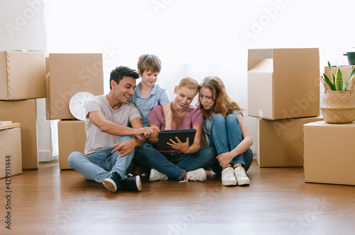 Happy caucasian family with teenage daughter and younger brother sitting on floor among cardboard boxes have fun using modern tablet buying furniture into new house.E-commerce retail services concept.
