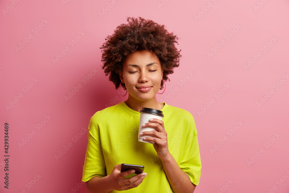 Pleased young curly Afro American woman closes eyes from smelling pleasant coffee aroma has relaxed face uses smartphone for chatting online dreessed in casual wear isolated over pink background
