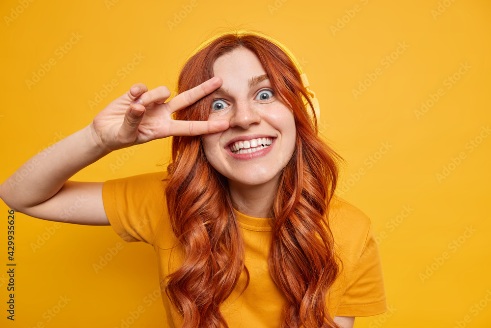 Happy cheerful female teenager smiles toothily has natural ginger hair makes peace gesture over eye listens music bia headphones wears casual t shirt poses against yellow background has fun. Happy cheerful female teenager smiles toothily has natural ginger hair makes peace gesture over eye listens music bia headphones wears casual t shirt poses against yellow background has fun.