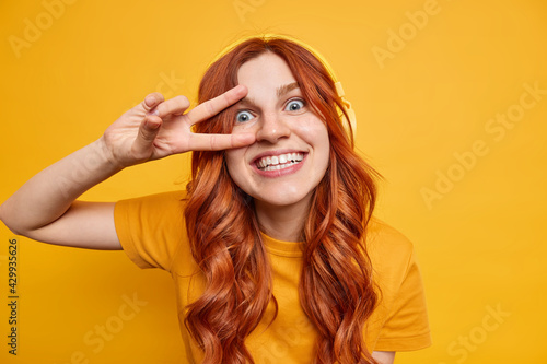 Happy cheerful female teenager smiles toothily has natural ginger hair makes peace gesture over eye listens music bia headphones wears casual t shirt poses against yellow background has fun.