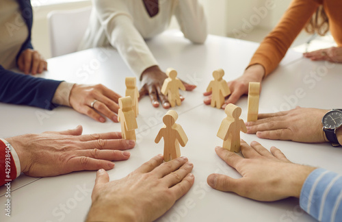 Team of multiracial business people and workmates sitting around white office table put little wooden human figures in circle as symbol of group, community, help, collaboration and teamwork