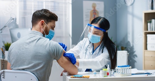 Close up portrait of pretty Asian female doctor in medical mask and face shield working in clinic cabinet during vaccination to handsome Caucasian male patient. Covid pandemic  vaccine cure concept