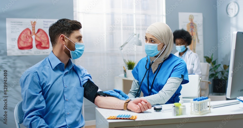 Pretty young Arab female nurse sitting in cabinet in hospital measuring blood tension to Caucasian man patient. Healthcare worker in medical center during covid-19 quarantine. Health concept