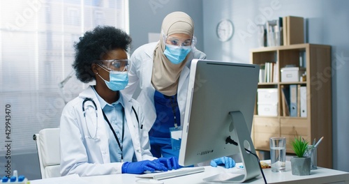 Portrait of mixed-race healthcare specialists working in cabinet in clinic. Arabic woman nurse speaking with African American young physician typing on computer browsing online. Hospital concept