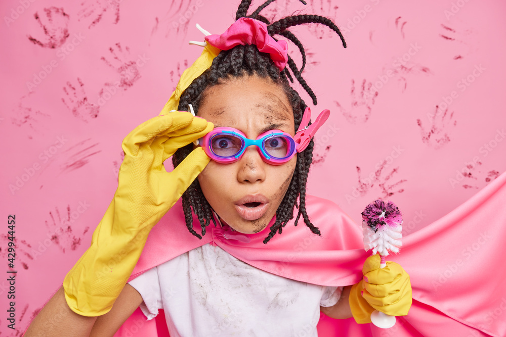 Everything should be perfect and clean. Surprised dirty ethnic woman wears goggles dressed like superhero holds toilet brush does quickly cleaning poses against pink background has lot of work